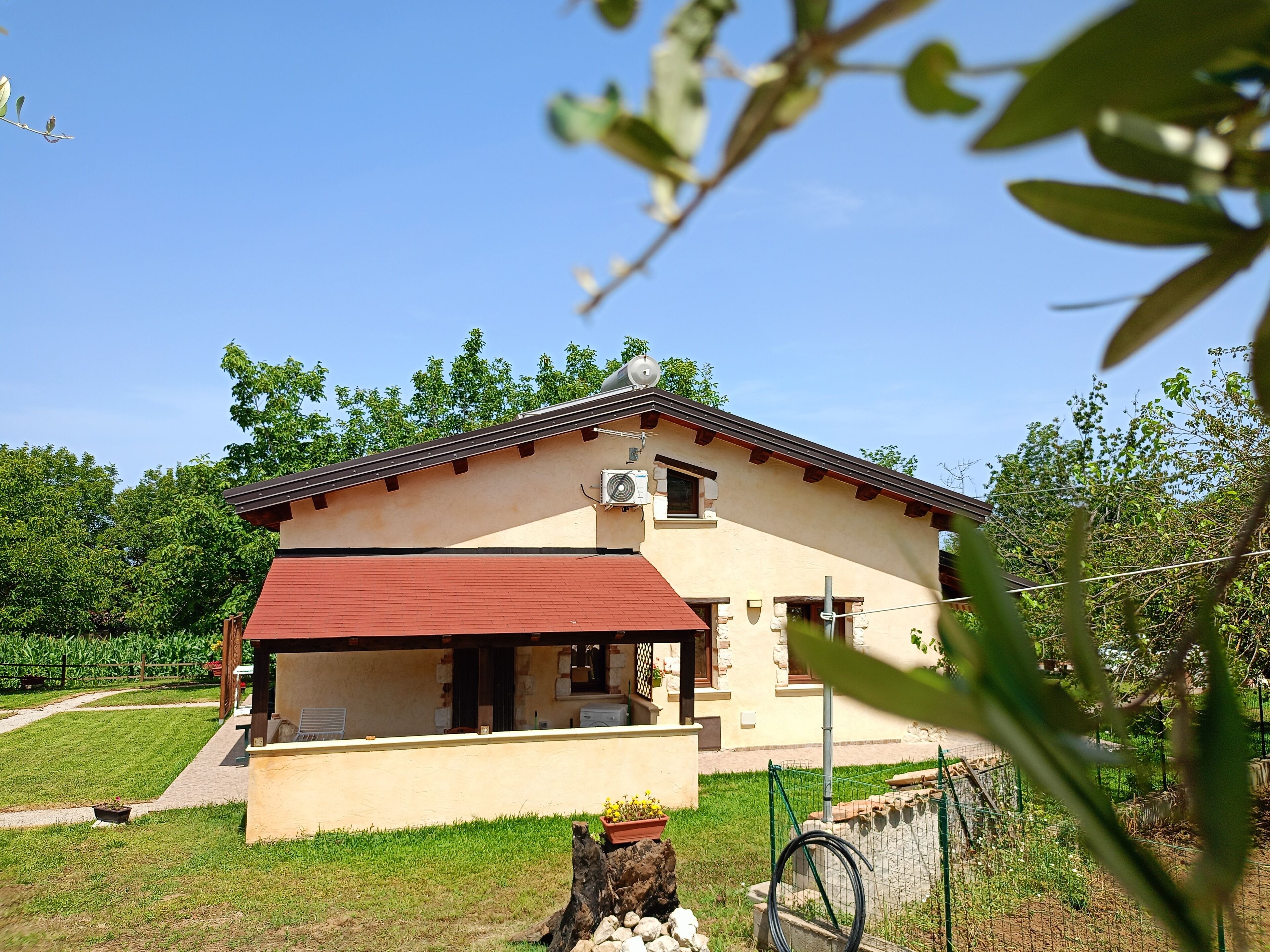 A two-story house with a red roof and beige walls, surrounded by greenery and a clear sky.