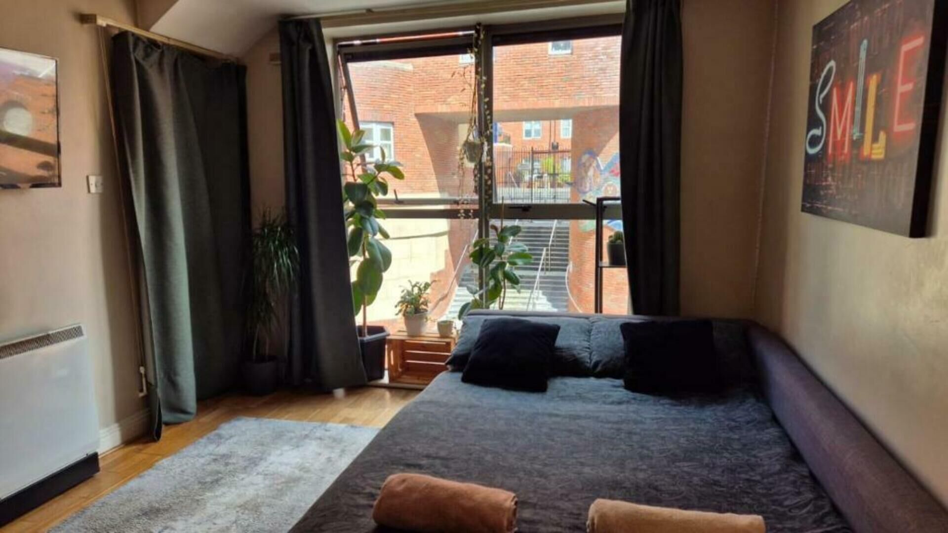 Un dormitorio con una cama grande, una ventana con cortinas, una planta en maceta y vistas a un edificio de ladrillo.