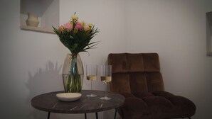 A round table with a vase of flowers and two glasses of white wine, a brown upholstered chair, and a small shelf with a vase.