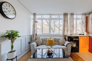 A living room with a sofa, a coffee table, a potted plant, and a clock on the wall. - Quaint 1BD Flat with Balcony in Maida Vale (Greater London)