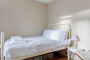 Una cama bien hecha con sábanas y almohadas blancas, una mesita de noche con lámpara, y una silla.