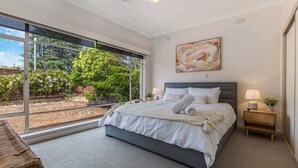 Quarto com uma janela grande, cama com roupa de cama branca, cabeceira cinza e um quadro na parede.