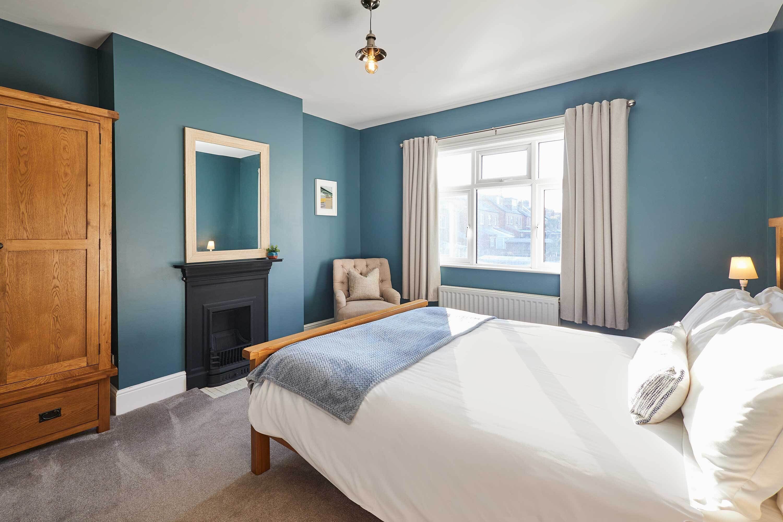 A bedroom with a bed, a wooden wardrobe, a fireplace, a chair, and a window with curtains.