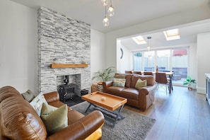 A modern living room with a stone fireplace, leather sofas, a wooden coffee table, and a dining area with natural light from skylights.