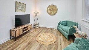 Un salon moderne avec un meuble de rangement en bois pour l’équipement audiovisuel, un téléviseur à écran plat, un canapé vert, un tapis rond tissé et une horloge murale.