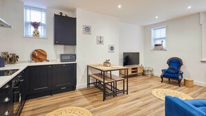 Una cocina moderna con armarios oscuros, una encimera de madera y un microondas integrado. Hay una zona de comedor con una mesa de madera y bancos, además de un sillón azul. La habitación tiene suelo de madera y alfombras tejidas decorativas.