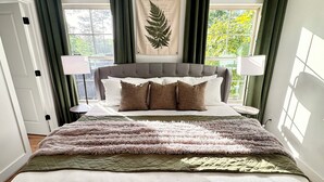 A bedroom with a large bed, green curtains, a botanical print on the wall, and a view of greenery outside.