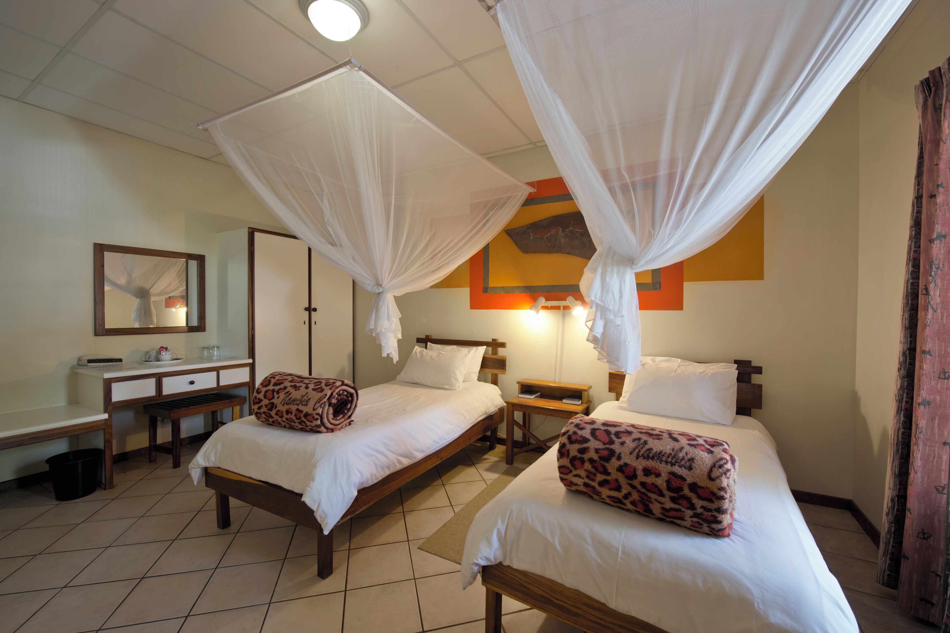Quarto de hotel com duas camas, cada uma com dossel, uma escrivaninha com cadeira, mesa de cabeceira e espelho.