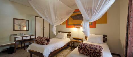 Quarto de hotel com duas camas, cada uma com dossel, uma escrivaninha com cadeira, mesa de cabeceira e espelho.