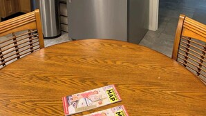 Una cocina con armarios de madera, un refrigerador de acero inoxidable, un microondas y un bote de basura. Hay una mesa de comedor con sillas y una revista encima.