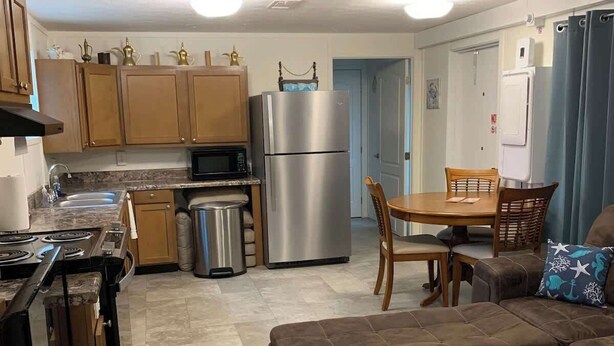 Un espacio habitable compacto con kitchenette, comedor y zona de estar con sofá y reposapiés.