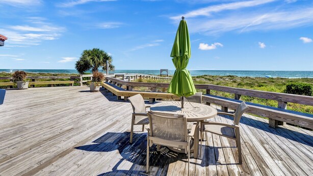 Una terraza de madera con una mesa y sillas, con vista a una playa bajo un cielo despejado.
