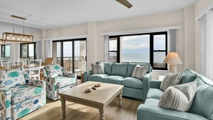 Un amplio salón con un gran ventanal que ofrece vistas al océano, un ventilador de techo y una zona de comedor con mobiliario blanco.