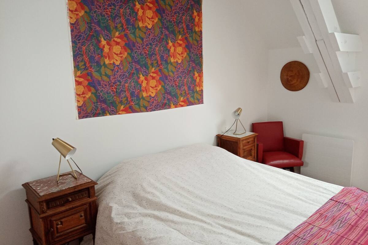 A bedroom with a bed, a nightstand, a red chair, a wall-mounted lamp, and a colorful floral tapestry.