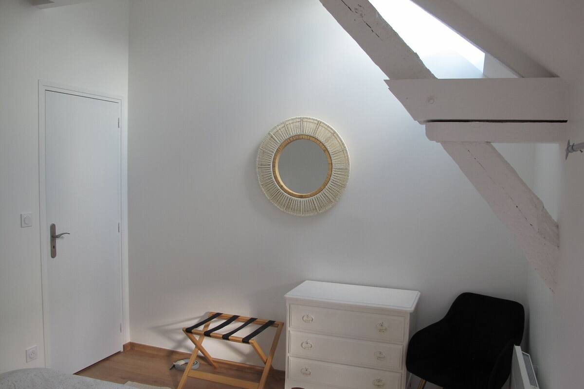 A bedroom with a white dresser, a black chair, a wooden drying rack, and a round mirror on the wall.