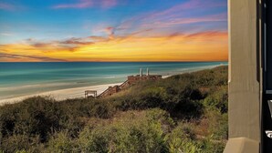 Vista costera con una escalera de madera que conduce a una playa, cielo despejado y una estatua sobre una plataforma.