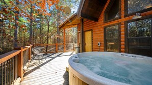 A wooden deck with a hot tub, a door, and a window overlooking a forest. - Burnt Whiskey! Beautiful Rustic Couples Cabin. (Broken Bow)