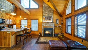 A cabin with a stone fireplace, wooden walls, a kitchen area, and a seating area with leather ottomans. - Burnt Whiskey! Beautiful Rustic Couples Cabin. (Broken Bow)