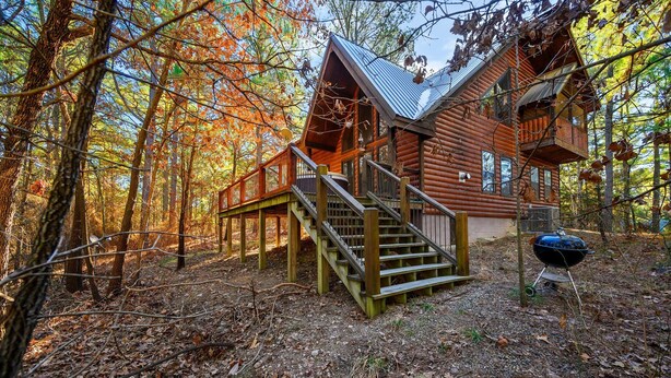 A log cabin with a wooden staircase leading to a deck, surrounded by a forest. - Burnt Whiskey! Beautiful Rustic Couples Cabin. (Broken Bow)