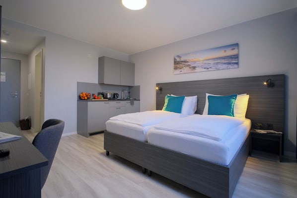 A modern hotel room with a double bed, a desk, a kitchenette, and a painting of a beach scene. - Hotel Kiel by MELIO (Melsdorf)