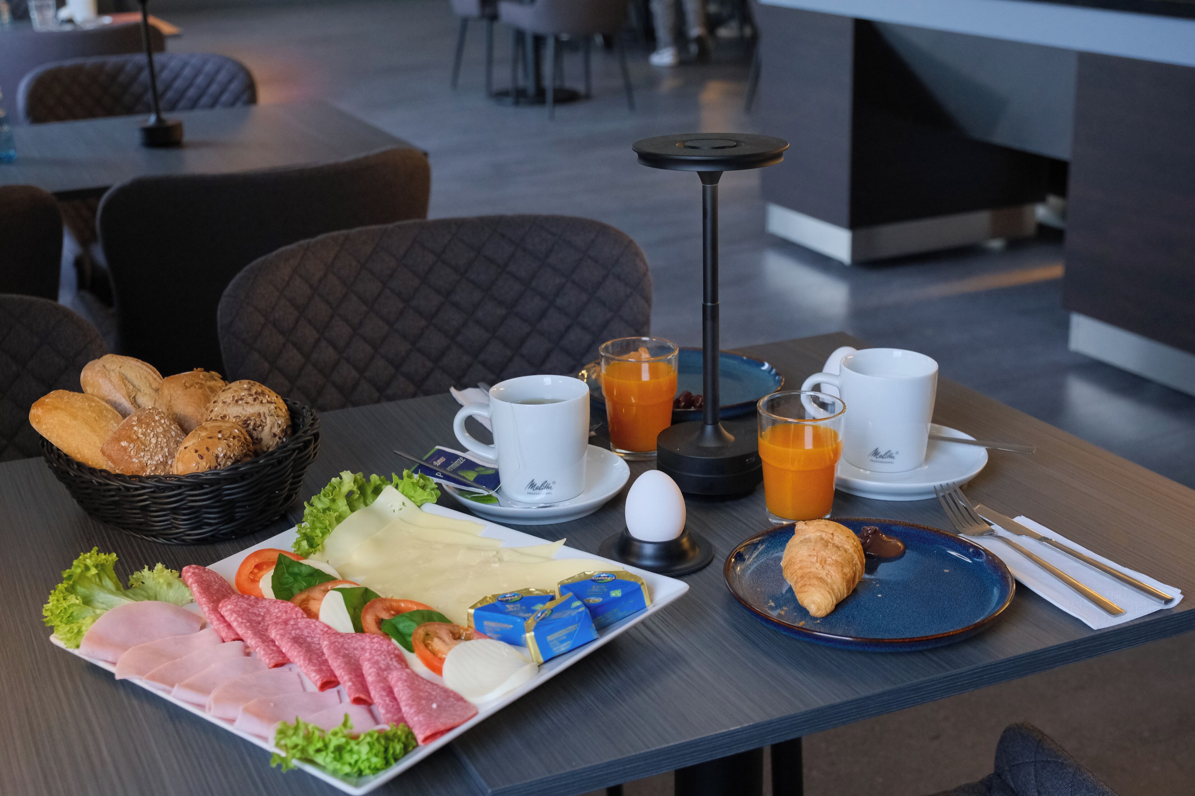 Een ontbijttafel met croissants, kaas, ham en brood. Twee koffiekopjes en een glas sinaasappelsap staan erbij.