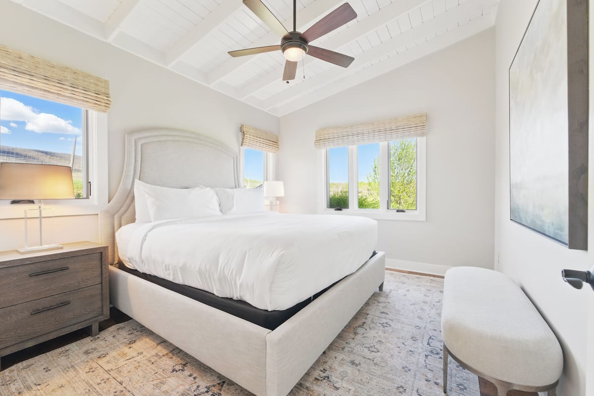 A bedroom with a large bed, a ceiling fan, and two windows with blinds.