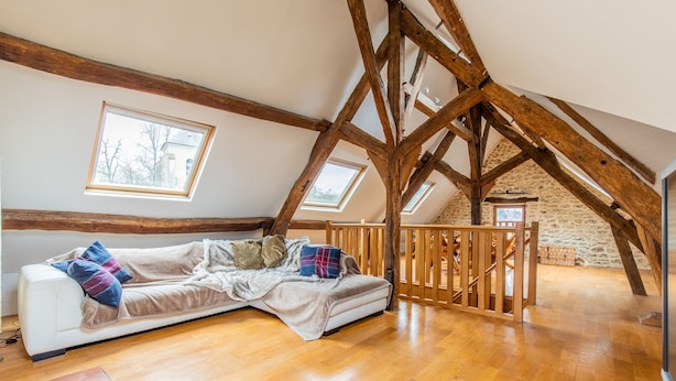 Ein gemütliches Schlafzimmer im Dachgeschoss mit schrägen Wänden, Holzbalken, einem weißen Sofa und zwei Dachfenstern.
