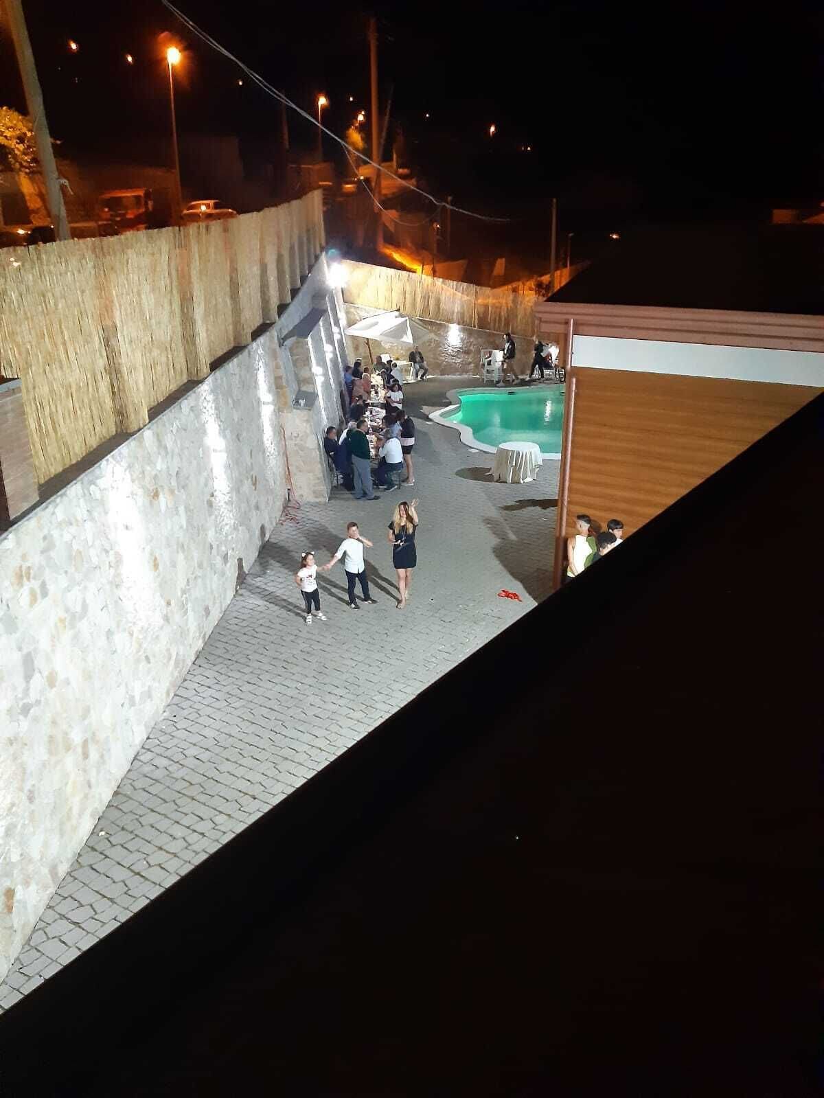Uma área externa à noite com piscina, espaço coberto para sentar e um caminho pavimentado.