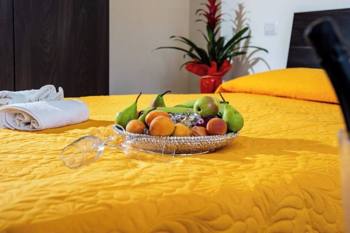 Un lit avec une couvre-lit jaune, un panier de fruits posé sur le lit, une serviette roulée et une plante en pot à l’arrière-plan.