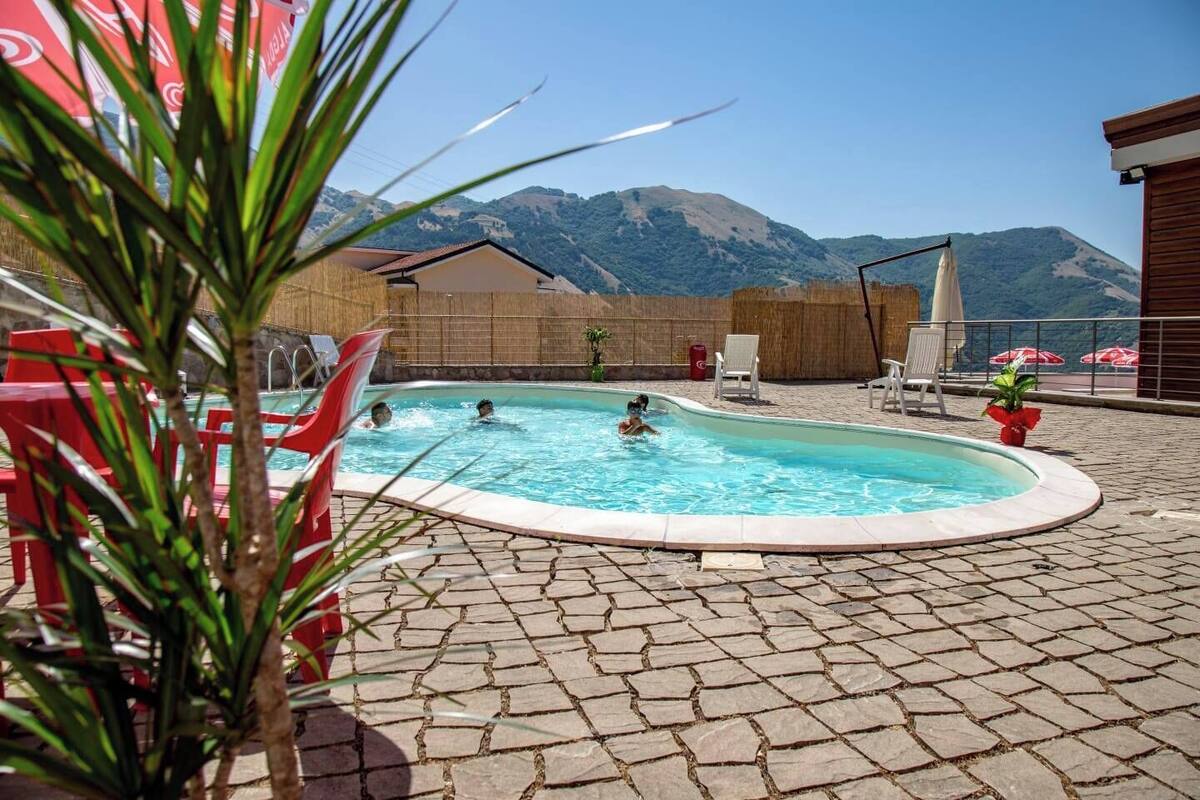 Une piscine où se baignent des personnes, entourée d’une terrasse en pierre, avec des montagnes en arrière-plan.