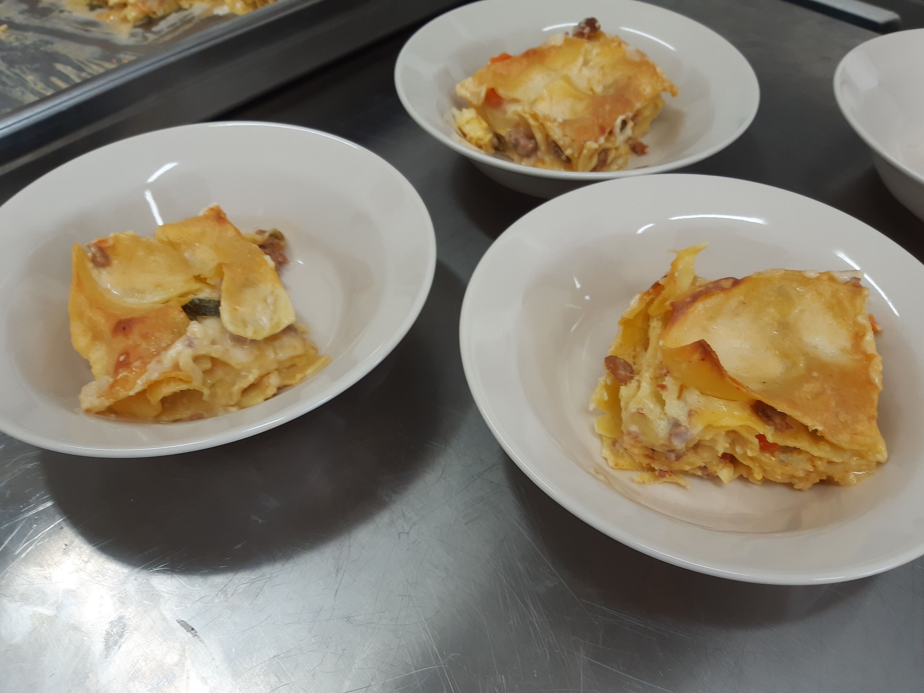 Four white plates with a baked dish, possibly lasagna, on a stainless steel surface.