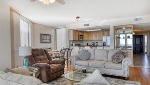 A spacious living room with a brown armchair, a grey sofa, and a glass coffee table. There is a dining area with a table and chairs, and a kitchen with wooden cabinets and a microwave. - One Ocean Place 1105- Three Bedroom, Three Bath Oceanfront Condo (Garden City Beach)