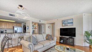 A living room with a sofa, a glass coffee table, a TV, and a kitchen area with a fridge and bar stools. - One Ocean Place 1105- Three Bedroom, Three Bath Oceanfront Condo (Garden City Beach)