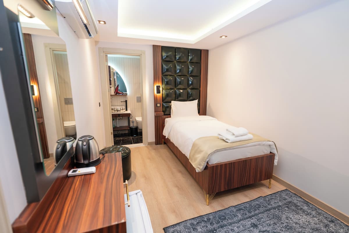 A hotel room with a wooden bed, a desk with a kettle, a mirror, and a small bathroom visible through an open door.