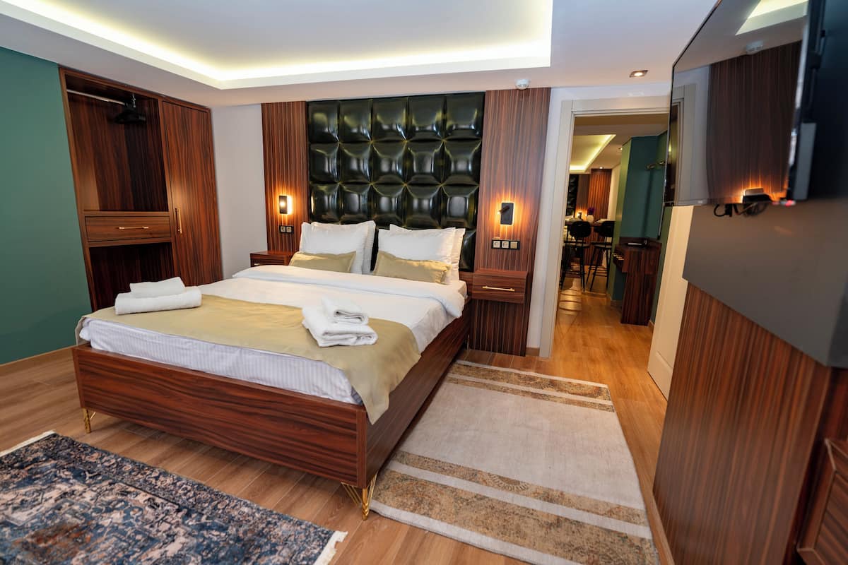 A modern bedroom with a large bed, wooden headboard, bedside tables with lamps, and a view into the dining area.