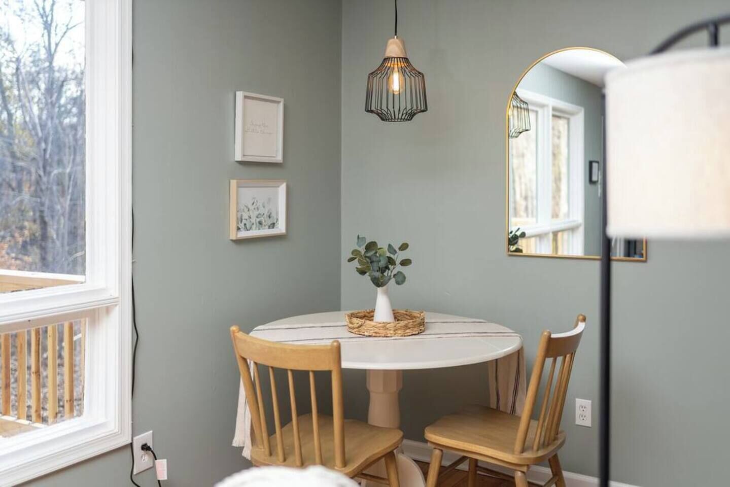 Un comedor con una mesa redonda, dos sillas de madera, un florero con hojas verdes y una lámpara colgante.