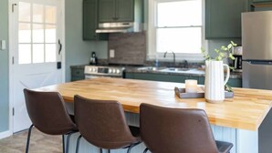 Une cuisine avec un îlot central en bois, des meubles de couleur vert foncé, un réfrigérateur en acier inoxydable et un vase blanc orné de verdure.