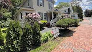 A two-story house with a porch, surrounded by lush greenery and flowering bushes. - Downtown Ogunquit Single home, 3 Bedroom (Ogunquit)