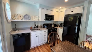 A compact kitchen with white cabinets, a black refrigerator, a microwave, and a stove. There is a wooden dining table and a chair. - Downtown Ogunquit Single home, 3 Bedroom (Ogunquit)