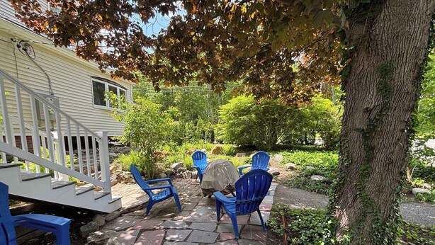 A covered patio with blue chairs and a table, surrounded by greenery and a tree. - Downtown Ogunquit Single home, 3 Bedroom (Ogunquit)
