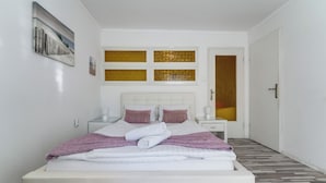 Ein ordentlich bezogenes Bett mit weißen Leinen und rosa Kissen, ein Nachttisch mit Lampe sowie ein Bild an der Wand.