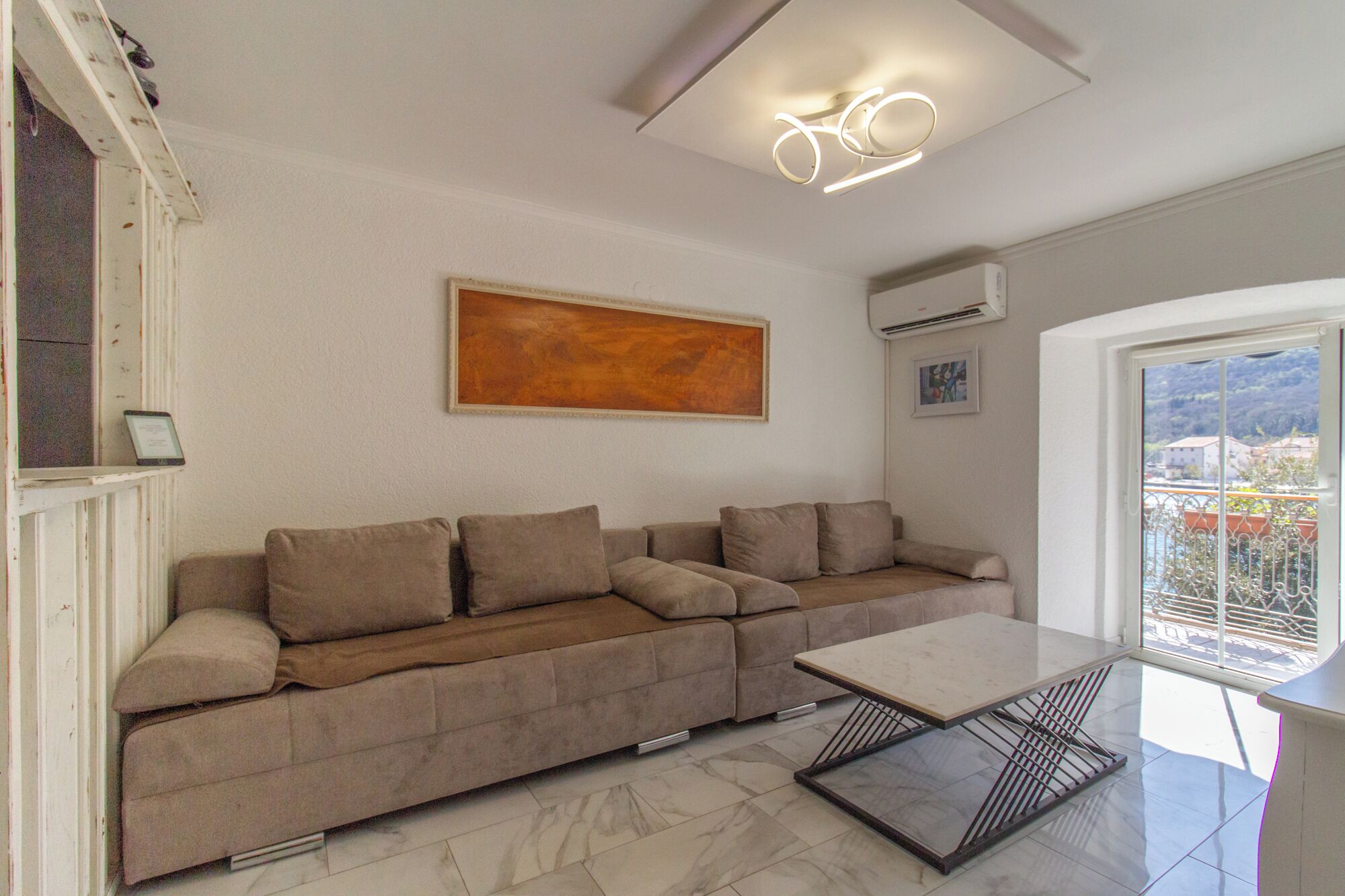 A modern living room with a beige sectional sofa, a glass coffee table, and a view of the outdoors through a large window.