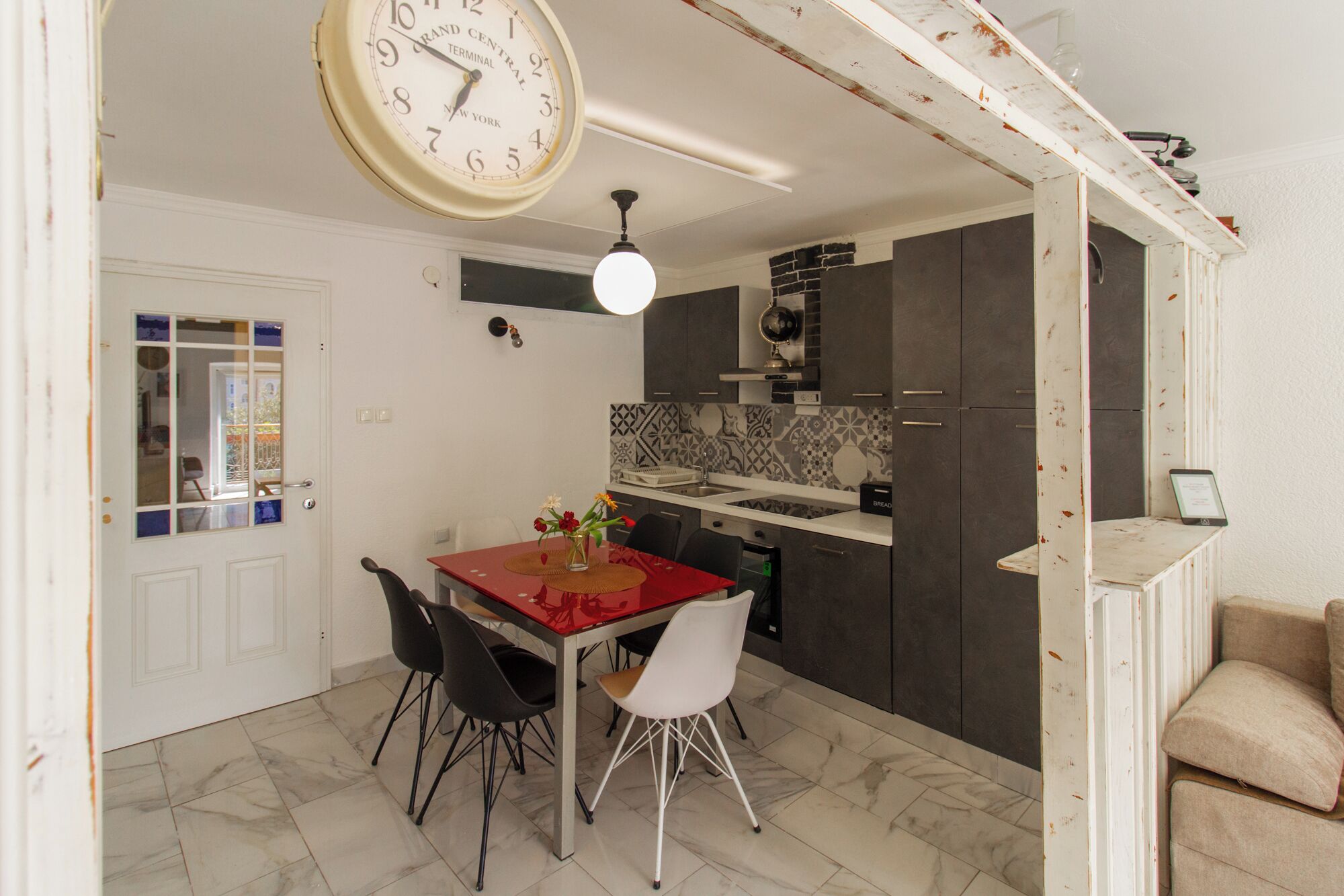 Un comedor con una mesa roja, sillas blancas y negras, un reloj y una cocina con armarios oscuros y salpicadero de azulejos.