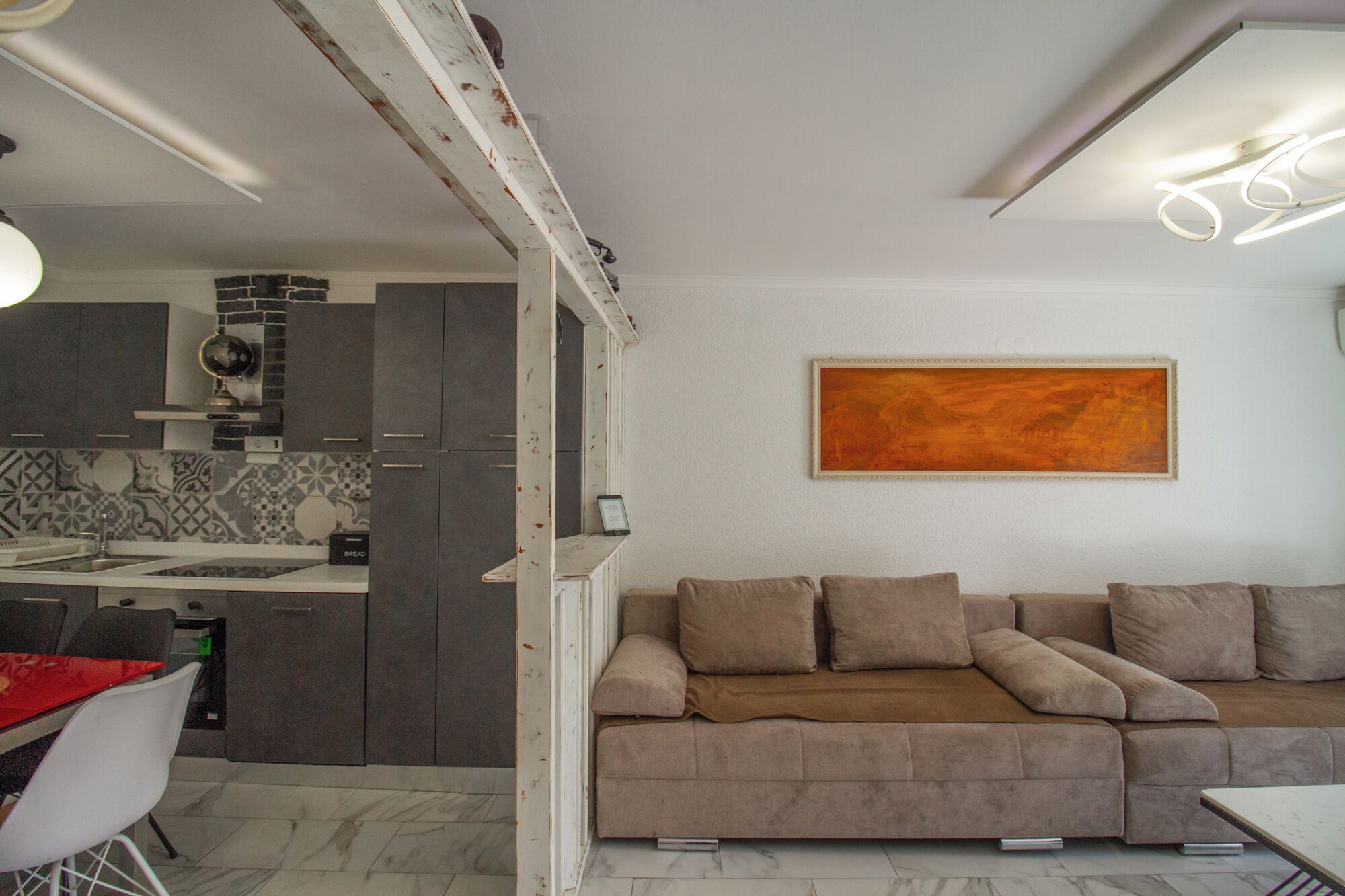 A modern living room with a beige sectional sofa, a kitchen with dark cabinets, and a large abstract painting on the wall.