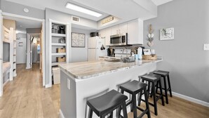 A modern kitchen with white cabinetry, a marble countertop, and a built-in microwave. There are three black bar stools and a small island with a sink. - Seacrest 905 by Liquid Life (Gulf Shores)