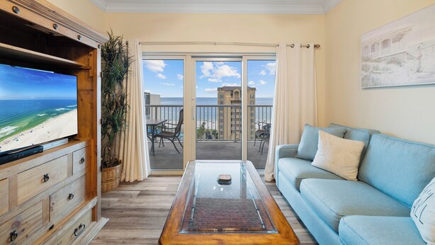A living room with a TV, a sofa, a glass coffee table, and a view of the beach and buildings through large windows. - Crystal Tower 807 by Liquid Life (Gulf Shores)