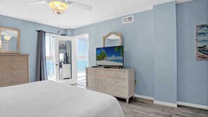 A bedroom with a large bed, a dresser, a television, and a view of the ocean through an open door.