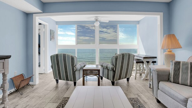 A room with a view of the ocean, two striped armchairs, a white coffee table, and a lamp.