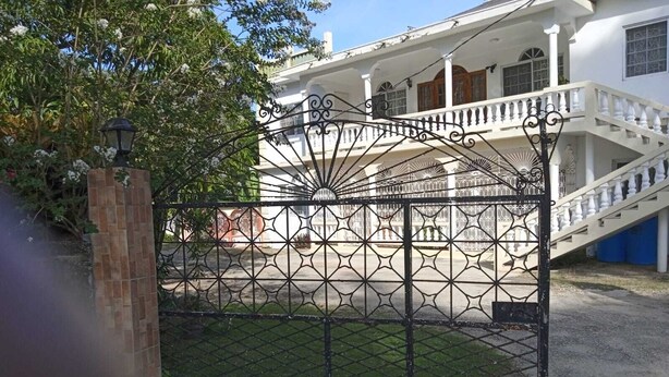 Ein verschlossener Eingang mit schmiedeeisernem Tor, einer Backsteinpfeiler und einem weißes Gebäude mit Balkon im Hintergrund.