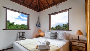 A bedroom with a bed, two bedside tables, a chair, and large windows with a view of greenery.